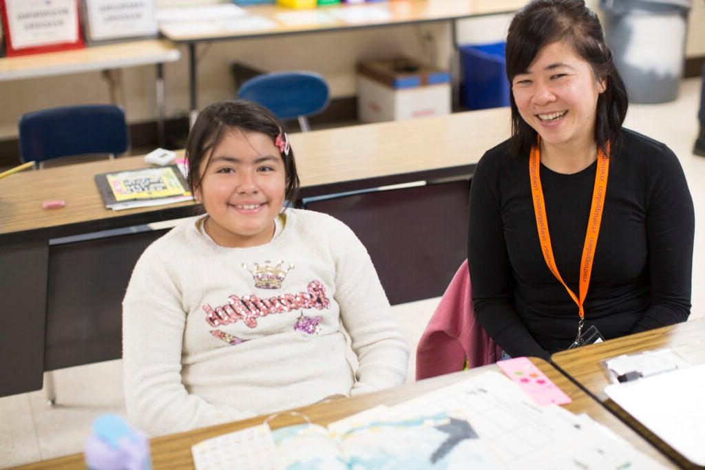Volunteer tutor and a student sitting at a desk in a classroom side-by-side smiling. 